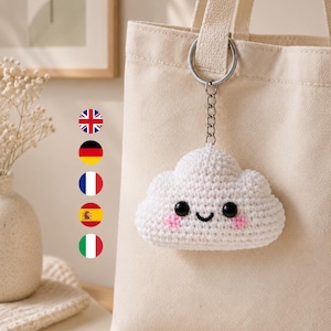 Può includere: Un portachiavi a forma di nuvola lavorato all'uncinetto bianco con occhi in plastica nera, un sorriso nero ricamato e guance rosa. La nuvola è fissata a un anello e una catena in metallo argentato, agganciati a una borsa in tela beige.