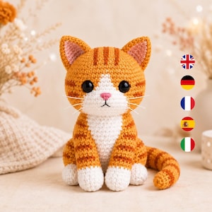 Könnte beinhalten: Ein gehäkeltes orange-weißes getigertes Katzenspielzeug mit dunkelbraunen Streifen, großen schwarzen Kunststoffaugen, einer kleinen rosa Nase und weißen Schnurrhaaren. Die Katze sitzt aufrecht mit seitlich gekringeltem Schwanz und weißen Pfoten.