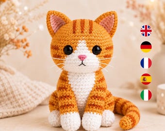 Könnte beinhalten: Ein gehäkeltes orange-weißes getigertes Katzenspielzeug mit dunkelbraunen Streifen, großen schwarzen Kunststoffaugen, einer kleinen rosa Nase und weißen Schnurrhaaren. Die Katze sitzt aufrecht mit seitlich gekringeltem Schwanz und weißen Pfoten.