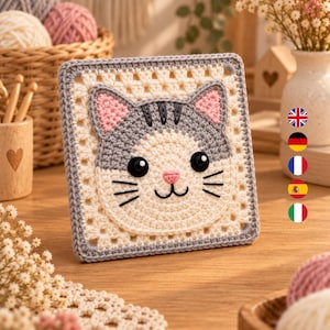 Könnte beinhalten: Ein quadratisches Häkelquadrat mit einem zentralen Katzengesicht-Motiv. Die Katze hat eine cremefarbene Schnauze, graue Ohren und Kopf, rosa Innenohren, schwarze Knopfaugen sowie aufgestickte schwarze Schnurrhaare und einen Mund.