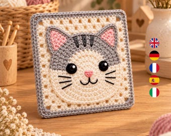 Könnte beinhalten: Ein quadratisches Häkelquadrat mit einem zentralen Katzengesicht-Motiv. Die Katze hat eine cremefarbene Schnauze, graue Ohren und Kopf, rosa Innenohren, schwarze Knopfaugen sowie aufgestickte schwarze Schnurrhaare und einen Mund.