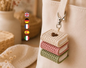 Könnte beinhalten: Eine cremefarbene Canvas-Einkaufstasche mit einem Häkel-Schlüsselanhänger. Der Anhänger besteht aus drei gestapelten Miniaturbüchern in Creme, Rosa und Salbeigrün, wobei auf dem obersten Buch ein kleines braunes Herz aufgestickt ist.