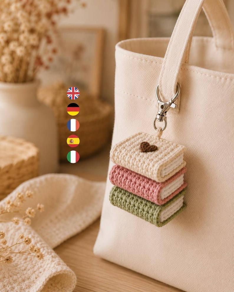 Puede incluir: Un bolso de tela color crema con un accesorio de llavero de ganchillo. El llavero consta de tres libros en miniatura apilados en color crema, rosa y verde salvia, con un peque&ntilde;o coraz&oacute;n marr&oacute;n bordado en la portada superior.