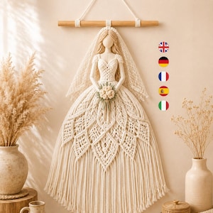Pode incluir: Uma peça de arte em macramé para parede que retrata uma figura num vestido de noiva. O cordão creme é atado num corpete detalhado e numa saia cheia e em camadas. Um véu a condizer pende de uma haste de madeira e segura um pequeno bouquet.