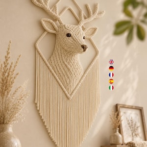 May include: A cream-coloured macrame wall hanging featuring a three-dimensional deer head sculpture. The deer is framed by a diamond-shaped rope structure with long, vertical fringe tassels extending downwards in a V-shape. The piece is off-white.