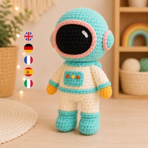 May include: A crocheted astronaut doll featuring a light blue helmet with a pink trim and a black visor. The doll has a white suit with light blue accents, orange hands, and a small rectangular chest panel with three colorful circular buttons.