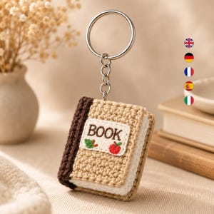 Puede incluir: Un llavero de ganchillo con forma de pequeño libro con cubierta de color tostado, lomo marrón oscuro y páginas blancas. Un parche rectangular blanco en la parte frontal presenta la palabra BOOK bordada en negro, junto a una hoja verde y una manzana roja.