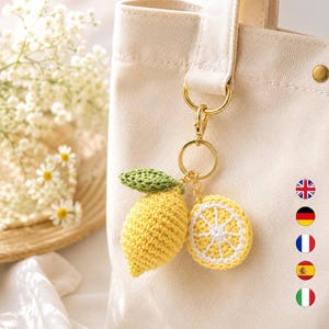 May include: A crochet lemon keyring featuring a yellow lemon fruit with a green leaf and a yellow lemon slice with white segments. The crochet pieces are attached to a gold-toned metal keyring and lobster clasp, which is clipped to a beige canvas tote bag.