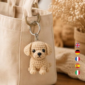 May include: A beige crochet dog keychain featuring a small puppy with black bead eyes and a black stitched nose. The keychain is attached to a silver metal ring, which is clipped onto the side of a light beige canvas tote bag with a handle.