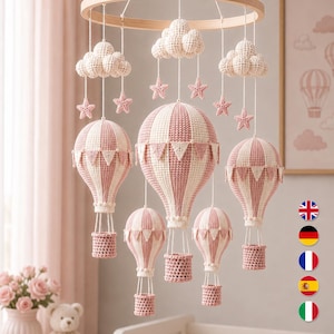 May include: A hanging mobile featuring five crocheted hot air balloons in alternating dusty pink and cream vertical stripes, accented with small triangular bunting. Three crocheted clouds and six small stars hang from a circular wooden frame.