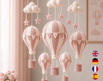 Könnte beinhalten: Ein hängendes Mobile mit fünf gehäkelten Heißluftballons in abwechselnden altrosa und cremefarbenen vertikalen Streifen, verziert mit kleinen dreieckigen Wimpeln. Drei gehäkelte Wolken und sechs kleine Sterne hängen an einem runden Holzrahmen.