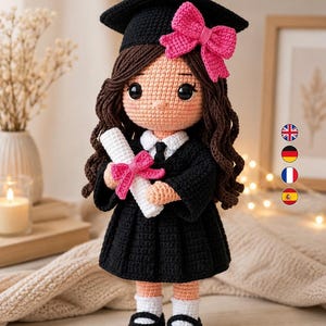 Puede incluir: Una muñeca de ganchillo vestida con toga y birrete negros, con cabello largo castaño, un lazo rosa brillante en el birrete y un diploma blanco atado con un lazo rosa a juego. La muñeca lleva una camisa blanca con cuello, calcetines blancos y zapatos negros.