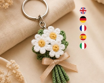 Könnte beinhalten: Ein Häkel-Schlüsselanhänger in Form eines kleinen Blumenstraußes aus drei weißen Gänseblümchen mit gelben Mitten und grünen Blättern. Die Stiele sind mit einer hellbeigen Satinschleife gebunden. Das Motiv ist an einem silberfarbenen Metallring befestigt.