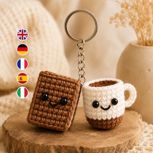 Puede incluir: Un llavero de ganchillo que presenta un libro rectangular marrón y una taza de café blanca con base marrón. Ambos artículos tienen ojos de cuentas negras y una sonrisa bordada. Las piezas están unidas por una anilla de metal plateado.