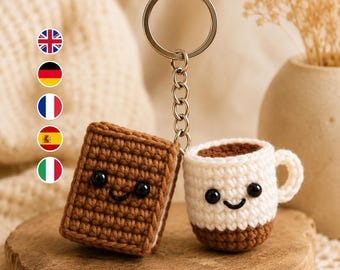 Könnte beinhalten: Ein Häkel-Schlüsselanhänger bestehend aus einem braunen rechteckigen Buch und einer weißen Kaffeetasse mit braunem Boden. Beide Teile haben schwarze Perlenaugen und ein gesticktes Lächeln. Sie sind mit einem silbernen Metallring verbunden.