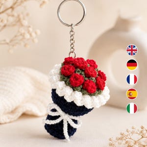 May include: A crochet keychain featuring a bouquet of small red roses with green stems, wrapped in a white ruffled border and a dark blue base tied with a white yarn bow. The bouquet is attached to a silver metal split ring and chain.