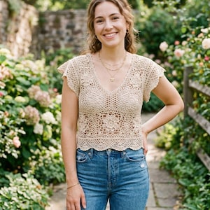 Könnte beinhalten: Beige gehäkeltes Oberteil mit kurzen Ärmeln, V-Ausschnitt und floralem Design. Das Oberteil wird mit einer blauen Jeans kombiniert. Das Bild wurde im Freien aufgenommen, mit einem Garten und einer Steinmauer im Hintergrund.
