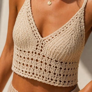 May include: A beige crochet crop top featuring a V-neckline and thin spaghetti straps. The garment is constructed with a combination of solid vertical ribbed stitching on the upper bodice and an openwork lattice pattern along the lower section.