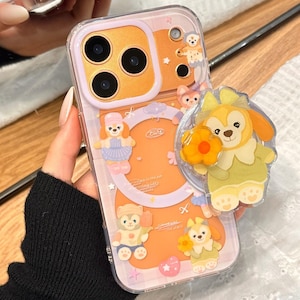 May include: Clear phone case with a cartoon character design. The case features a pop-up grip and a colorful design with cartoon characters in shades of orange, yellow, and pink. The case is designed to fit a smartphone.
