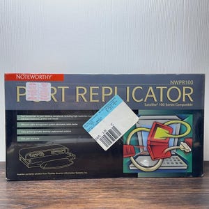 May include: A rectangular box for a Noteworthy NWPR100 Port Replicator, compatible with Satellite 100 Series. The box features a dark blue and black design with gold text, a line drawing of the device, and a graphic of a red laptop with cables.