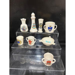 May include: A collection of eight white ceramic souvenir pieces, including a bell, a lighthouse figurine, a tall vase, a two-handled mug, a small cup, a two-handled cup, a small bowl with handles, and a two-handled mug, all featuring colorful heraldic crests.