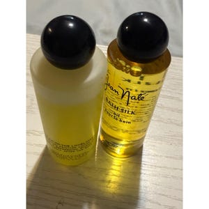 May include: Two cylindrical bottles with black spherical caps. The left bottle contains an opaque yellow lotion with text reading Tip-To-Toe Loveliness. The right bottle contains a clear golden bath silk oil labeled Jean Nate Bath Silk.
