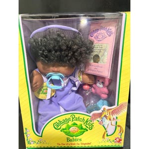 May include: A Cabbage Patch Kids baby doll in a box. The doll has dark skin, curly black hair, and is wearing a purple outfit and a pacifier. The box includes a birth certificate and adoption papers.
