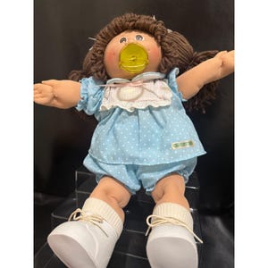 Könnte beinhalten: Eine Vintage Cabbage Patch Kids Puppe mit braunen Wollhaaren in Zöpfen, die ein hellblaues Kleid mit Punkten und passende Shorts trägt. Die Puppe hat einen gelben Schnuller und weiße Schuhe.