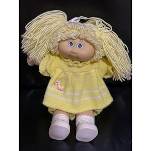 May include: A vintage Cabbage Patch Kid doll with blonde yarn hair styled in pigtails. The doll wears a yellow knit dress with a duck applique and white stripes, matching bloomers, and white shoes. The doll has blue eyes and a rosy complexion.