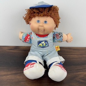 May include: A Cabbage Patch Kids doll with curly brown hair and blue eyes, wearing a blue and white striped cap, matching striped overalls with a colourful vehicle print, and red and white trainers. A small yellow tag is attached to the side.