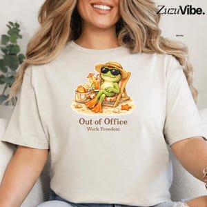 Può includere: T-shirt beige con un cartone animato di una rana con occhiali da sole e un cappello di paglia, che si rilassa su una sedia a sdraio con una bevanda. Il testo "Out of Office Work Freedom" è stampato sotto.