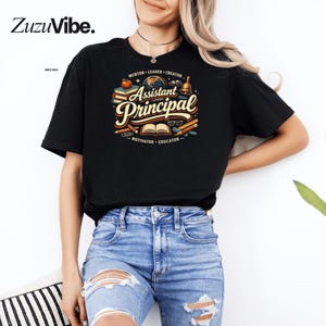 Può includere: T-shirt nero con un design grafico colorato per un assistente preside. Il design include le parole "Assistant Principal" in un font decorativo, circondate da libri, una mela, una campana e matite. La maglietta è abbinata a jeans blu.