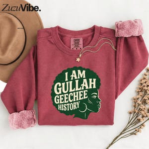 May include: A maroon sweatshirt with a green and white graphic that reads "I AM GULLAH GEECHEE HISTORY" and a profile of a woman. The sweatshirt has a fleece lining on the cuffs. A gold necklace with a star pendant is draped across the sweatshirt.