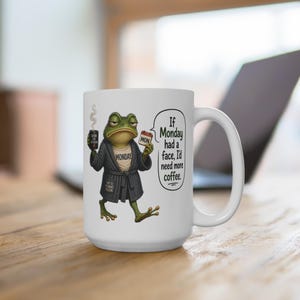 以下が含まれることがあります： 白セラミックコーヒーマグ。不機嫌そうなカエルのイラストが特徴です。カエルはバスローブを着てコーヒーを持っており、「If Monday had a face, I'd need more coffee」というテキストが書かれています。カエルはコーヒーカップと「MON」カードを持っています。