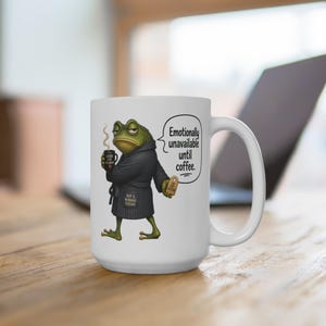 以下が含まれることがあります： 白いセラミックコーヒーマグ。ローブを着てコーヒーカップを持った漫画のカエルが描かれています。マグには「Emotionally unavailable until coffee」の文字が書かれています。カエルは緑色で不機嫌そうな表情をしています。