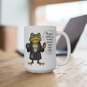 May include: White ceramic coffee mug featuring a cartoon frog in a robe, holding coffee and a donut. The frog has a grumpy expression and the mug has the text "Oh great, another day of making miracles happen before coffee."