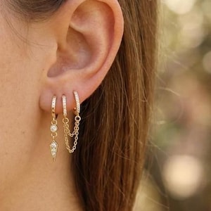 May include: Gold-toned earrings with sparkling accents are displayed on an ear. The earrings include small hoops, a chain, and a dangling geometric shape. The jewelry is set against a blurred background.
