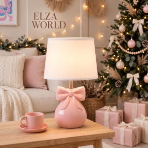 Puede incluir: Una lámpara de mesa con una base de cerámica rosa claro brillante en forma de bolsa redondeada rematada con un gran lazo. La lámpara cuenta con una pantalla de tela blanca y un vástago metálico. El texto ELZA WORLD es visible arriba a la izquierda.