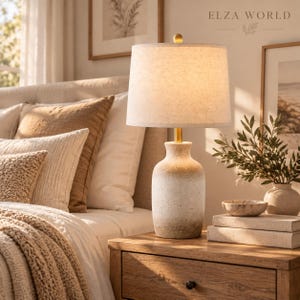 May include: A table lamp with a textured, off-white and tan ombre ceramic base and a cylindrical fabric shade. The lamp sits on a wooden nightstand next to a stack of two books, a small bowl, and a potted plant with green leaves.
