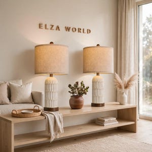 May include: Two table lamps with cylindrical white textured bases and dark wood accents, topped with beige fabric drum shades. The lamps are placed on a light wood console table, with the text ELZA WORLD mounted on the wall above them.