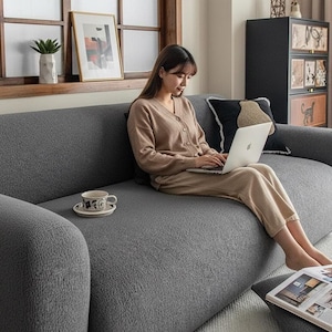 May include: A contemporary grey sofa with curved arms and a matching footstool. A person is using a laptop while seated on the sofa. A small wooden side table with a potted plant and a teacup are nearby. The room has a window and a decorative cabinet.