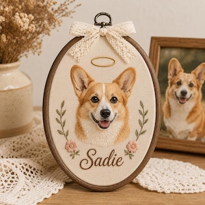 Puede incluir: Un bastidor de bordado ovalado de madera que contiene un retrato en tela de un perro Corgi con un halo dorado sobre su cabeza. El perro está flanqueado por dos tallos florales bordados y el nombre Sadie está cosido en cursiva marrón.