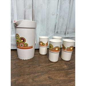 May include: A white pitcher and four matching tumblers. The pitcher and tumblers have a retro design with green, brown, and orange circular patterns. The pitcher has a pink interior and a handle.