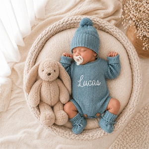 May include: A sleeping baby wearing a blue knit outfit, hat, and booties, with the name "Lucas" embroidered on the chest. A plush bunny toy sits beside the baby in a woven basket. The baby has a pacifier in its mouth.