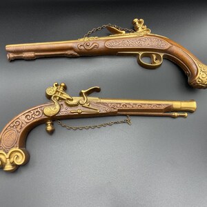 May include: Two antique replica flintlock pistols. The pistols have a brown wooden handle and barrel with ornate gold-colored metal accents. The pistols are detailed with engraved patterns and connected by a small chain.