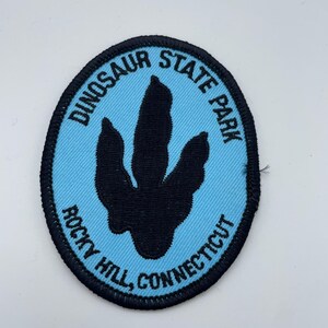 May include: An oval patch featuring a black dinosaur footprint on a light blue background. The patch is embroidered with the words "Dinosaur State Park" and "Rocky Hill, Connecticut" in black lettering, surrounded by a black border.