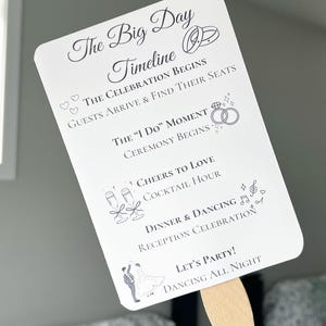 May include: A white wedding fan with a wooden handle, featuring a timeline of events. The text includes "The Big Day Timeline," "The Celebration Begins," "The 'I Do' Moment," and "Let's Party!" with decorative illustrations.