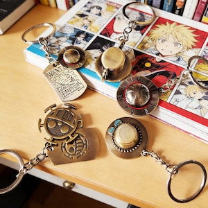 Może przedstawiać: Kolekcja breloków w kolorze brązu o różnych wzorach. Jeden brelok ma nieśmiertelnik z napisem "WANT" i ilustracją postaci. Inne breloki mają wzory kapeluszy i emblematów. Breloki leżą na drewnianej powierzchni.