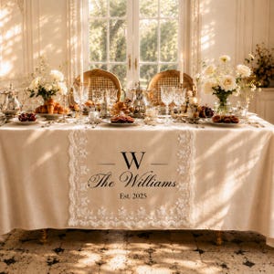 May include: A beige linen table runner with white lace trim, featuring a black printed monogram with the letter W, the name The Williams, and the text Est. 2025, centred on a dining table set with silver teapots, glassware, and floral arrangements.
