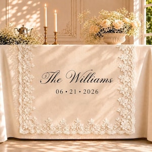 Op de afbeelding: Een beige tafelkleed met gedetailleerde witte kanten randen. In het midden staat de zwarte tekst The Williams en de datum 06 21 2026. De tafel is versierd met twee hoge kaarsen in gouden houders en een boeket met witte bloemen.